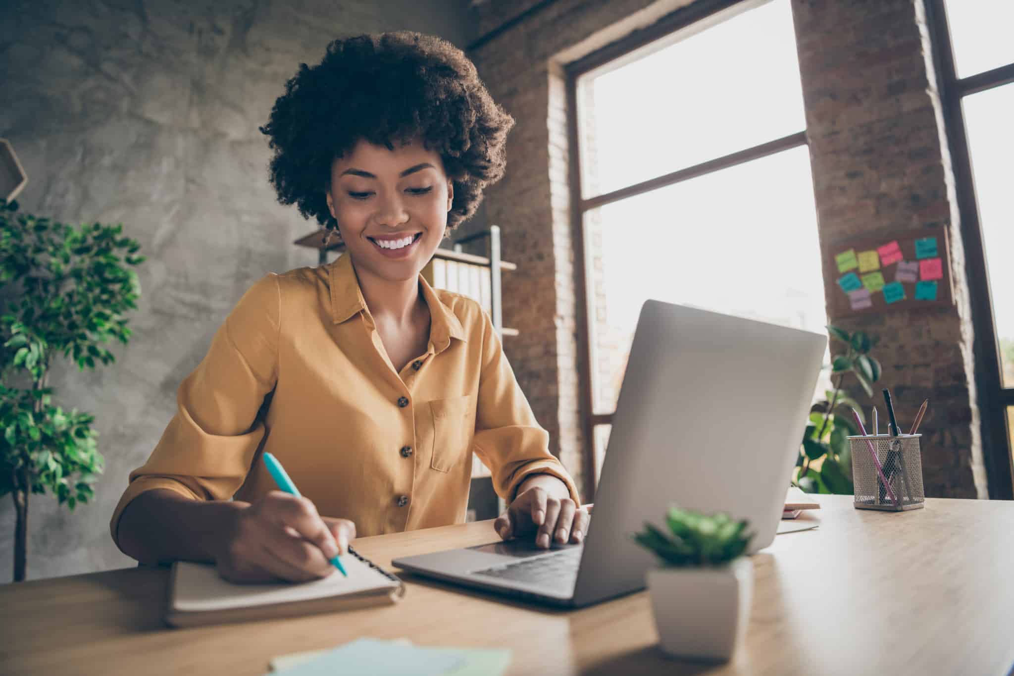 Cheerful female employee sitting at desktop with laptop noting down important information, company career development seminar